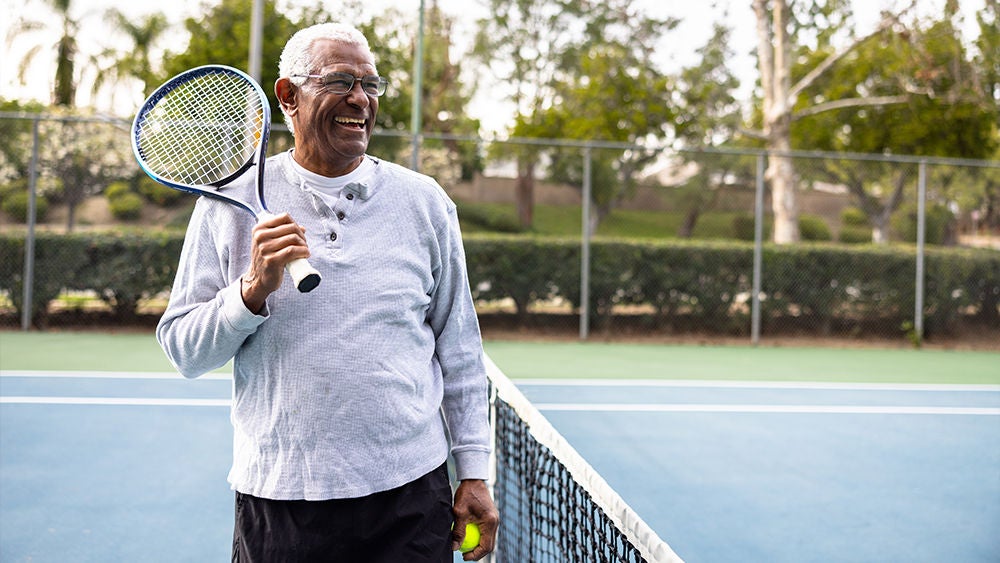 Ein älterer Mann steht lachend auf einem Tennisplatz und hält seinen Schläger auf die Schulter.